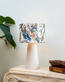 A table lamp with a cream-colored tapered base and a cylindrical fabric shade featuring a detailed bird and floral print, styled on a marble console table next to a green plant and books.