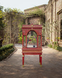 Red decorative bird cage in an outdoor setting with a building and greenery in the background