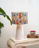 A table lamp with a cream-colored tapered base and a cylindrical drum shade featuring a colorful, detailed traditional Indian illustration pattern, styled on a marble console table next to a plant and a small bowl.