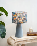 A table lamp with a textured light blue-gray cone base and a cylindrical drum shade featuring a multicolor floral and botanical pattern, styled on a marble console table next to a plant and a small Buddha head statue.