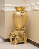 A decorative gold-toned vase with a textured finish and hanging ring, displayed on an elephant-shaped pedestal in a beige interior corner.