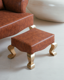 Brown leather footstool with gold legs next to a brown leather chair on a light-colored floor.