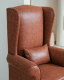 Brown leather wingback chair with a pillow against a neutral background