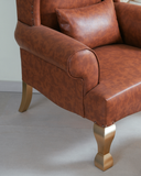 Brown leather armchair with gold legs on a light gray floor.