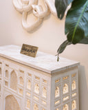 Decorative white cabinet with intricate carvings and a small sign on top, against a light wall.