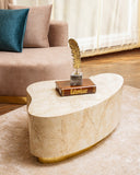 A wavey marble coffee table with a gold metal base, styled in a living room setting with a sofa, decorative book, and feather sculpture on top.