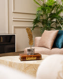 A decorative gold feather quill sculpture on a gray stone base, displayed on a brown book engraved with 'Kalamkaar', set on a marble coffee table in a modern living room with a beige sofa and green plant.