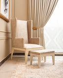 A beige upholstered wingback armchair with a matching ottoman, placed in a bright room with cream curtains, a patterned rug, and a beige wall.