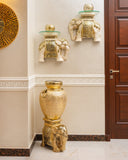 A decorative gold elephant-shaped pedestal stand holding a large textured gold urn, positioned in a hallway next to a dark wood door. Two smaller matching gold elephant wall shelves with glass tops are mounted higher on the beige wall.